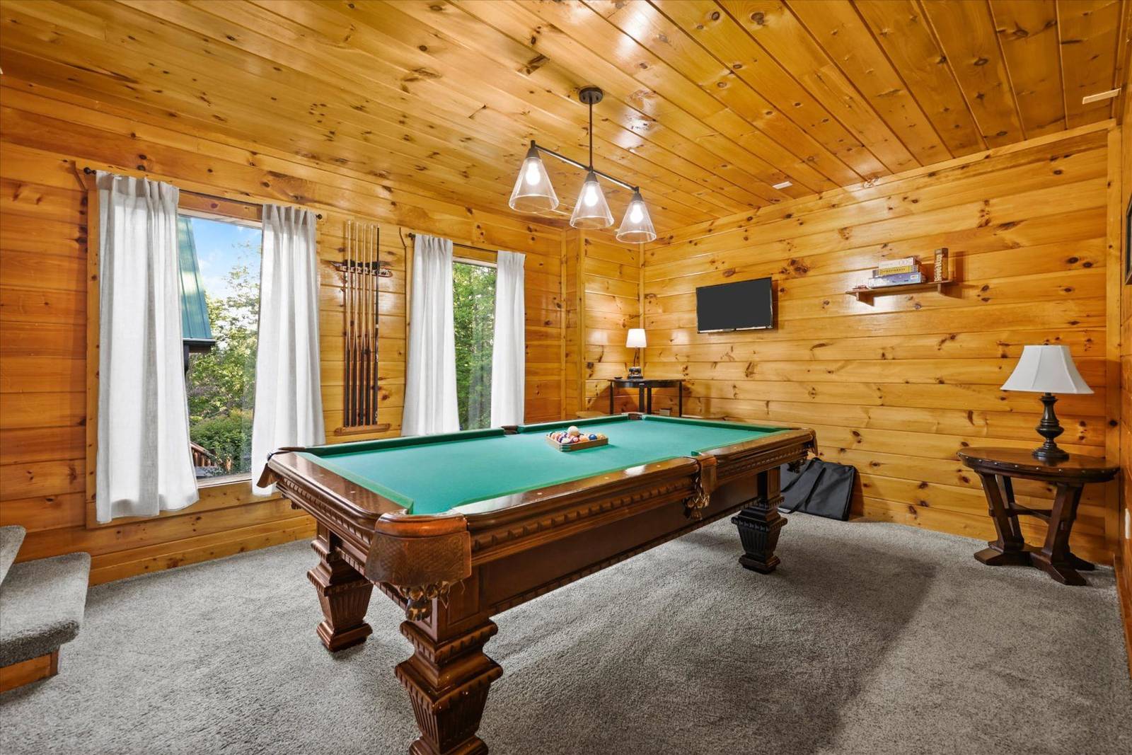 Rustic wooden game room with pool table, windows, TV, and cue rack