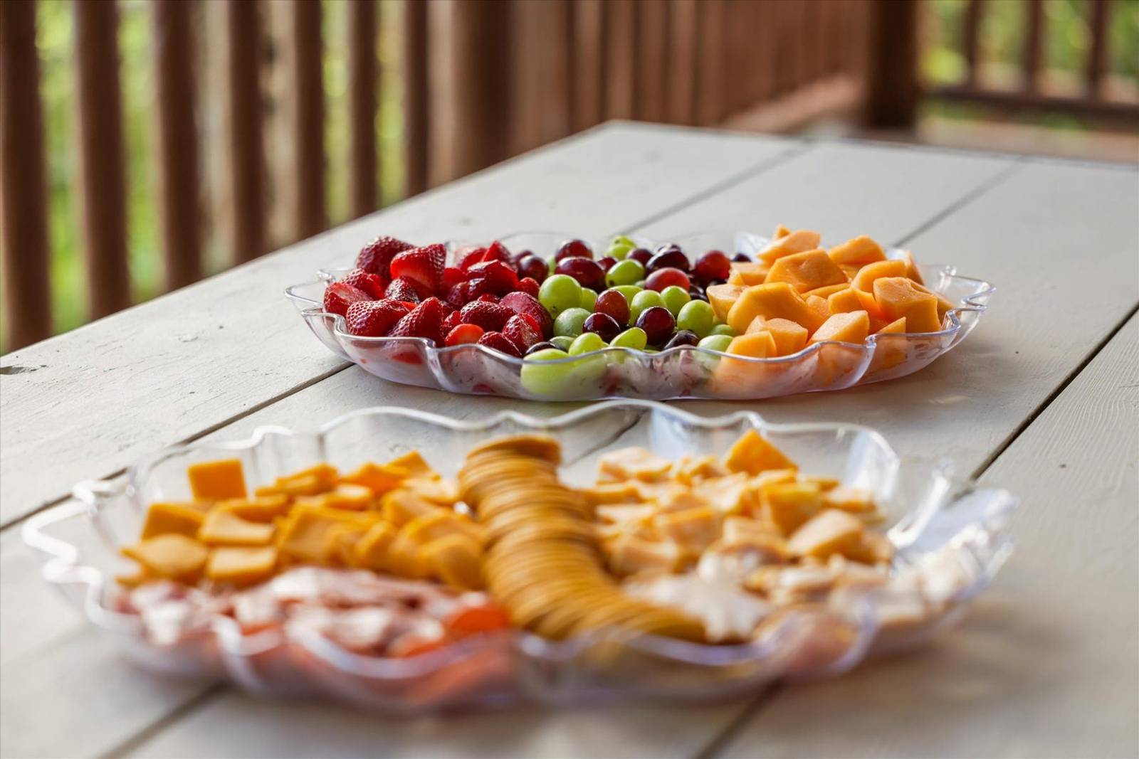 Two clear platters of fruit, cheese, and crackers on a wooden table
